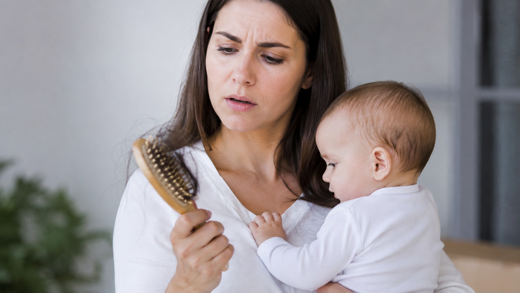 chute de cheveux post partum, jeune maman avec bébé tenant une brosse remplie de cheveux, perte capillaire après grossesse, intérieur lumineux, 2026, format 16/9