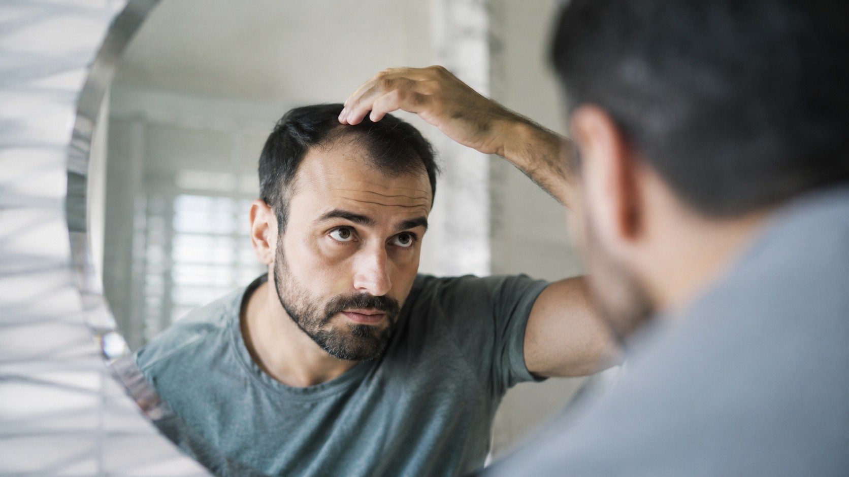 Homme observant une alopécie androgénétique au niveau de la ligne frontale dans un miroir de salle de bain, lumière naturelle douce, photo réaliste 2026 format 16/9