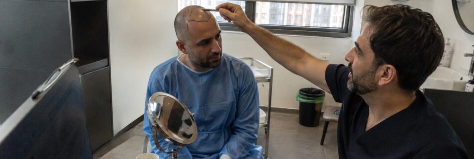 consultation préopératoire de greffe de cheveux en Turquie avec patient au crâne rasé portant une blouse médicale bleue, praticien traçant la ligne frontale au marqueur, miroir de contrôle sur bureau, cabinet capillaire moderne éclairé par lumière naturelle, photographie documentaire Canon EOS R6 grand-angle 24–28 mm, août 2026