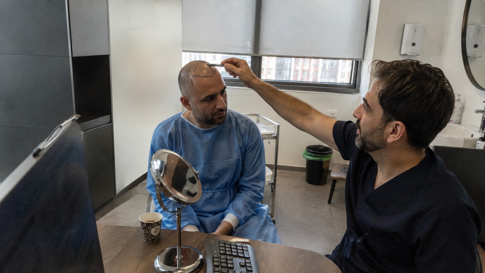 consultation préopératoire de greffe de cheveux en Turquie avec patient au crâne rasé portant une blouse médicale bleue, praticien traçant la ligne frontale au marqueur, miroir de contrôle sur bureau, cabinet capillaire moderne éclairé par lumière naturelle, photographie documentaire Canon EOS R6 grand-angle 24–28 mm, août 2026